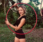 A woman in a black sleeveless crop top and shorts holds a large red hula hoop outdoors in a wooded setting.